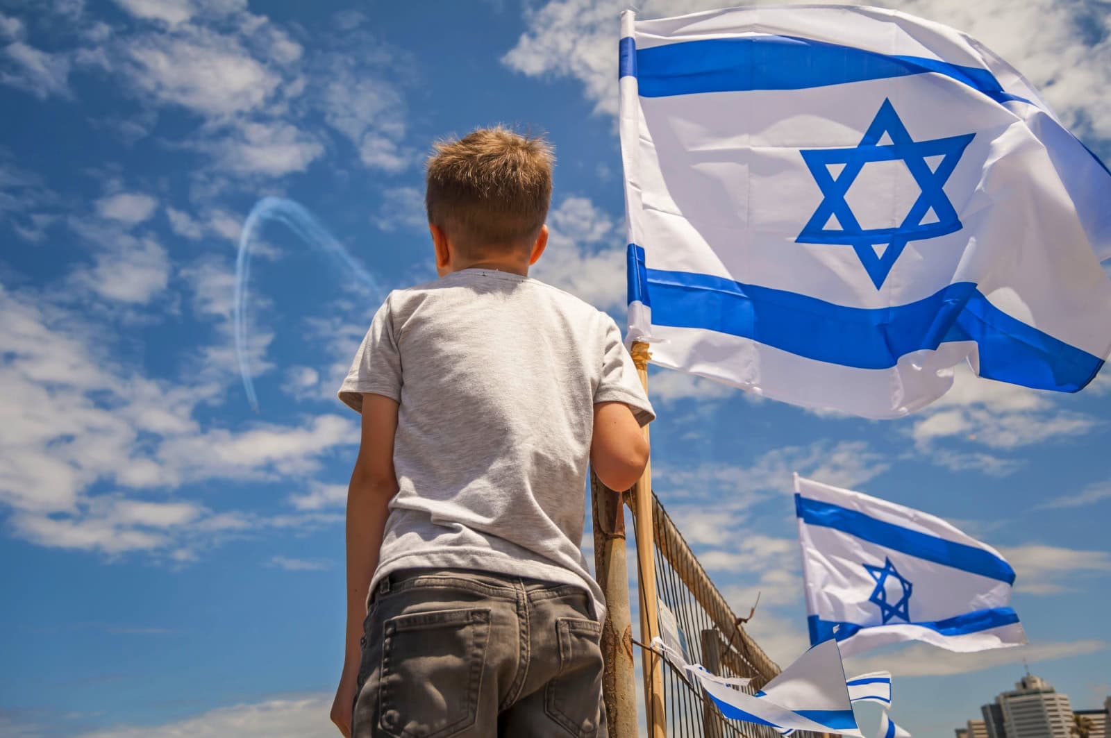 A young boy stands near a railing, facing away, from the camera to watch an air force show in the sky, with Israeli flags waving beside him under a partly cloudy blue sky.