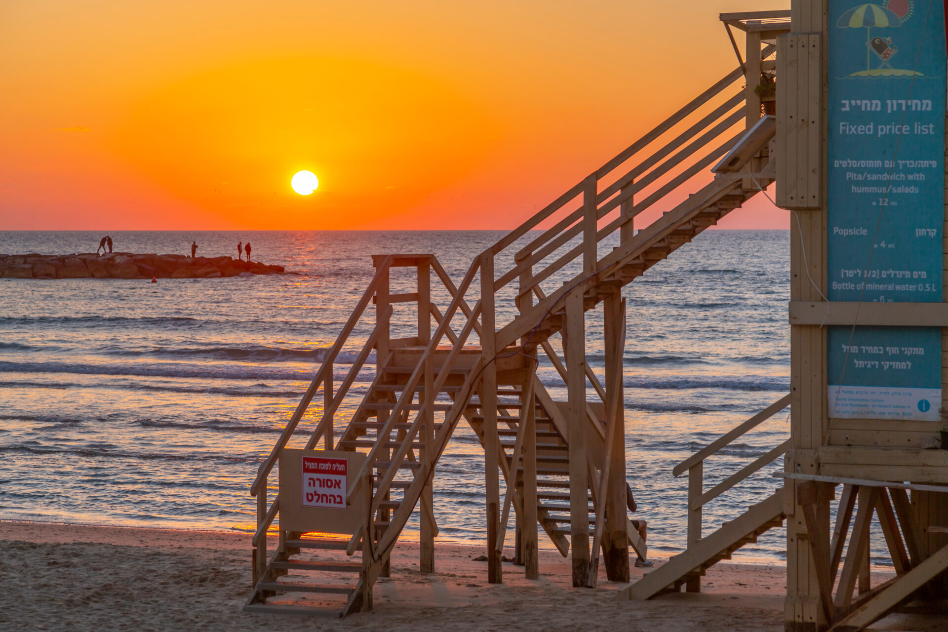 A lifeguard tower stands on Borgashov Beach at sunset, with a staircase leading down to the sand. The sun is setting over the horizon, casting an orange glow on the sea. A few people are visible on a distant rocky jetty. Signs in Hebrew are on the tower.