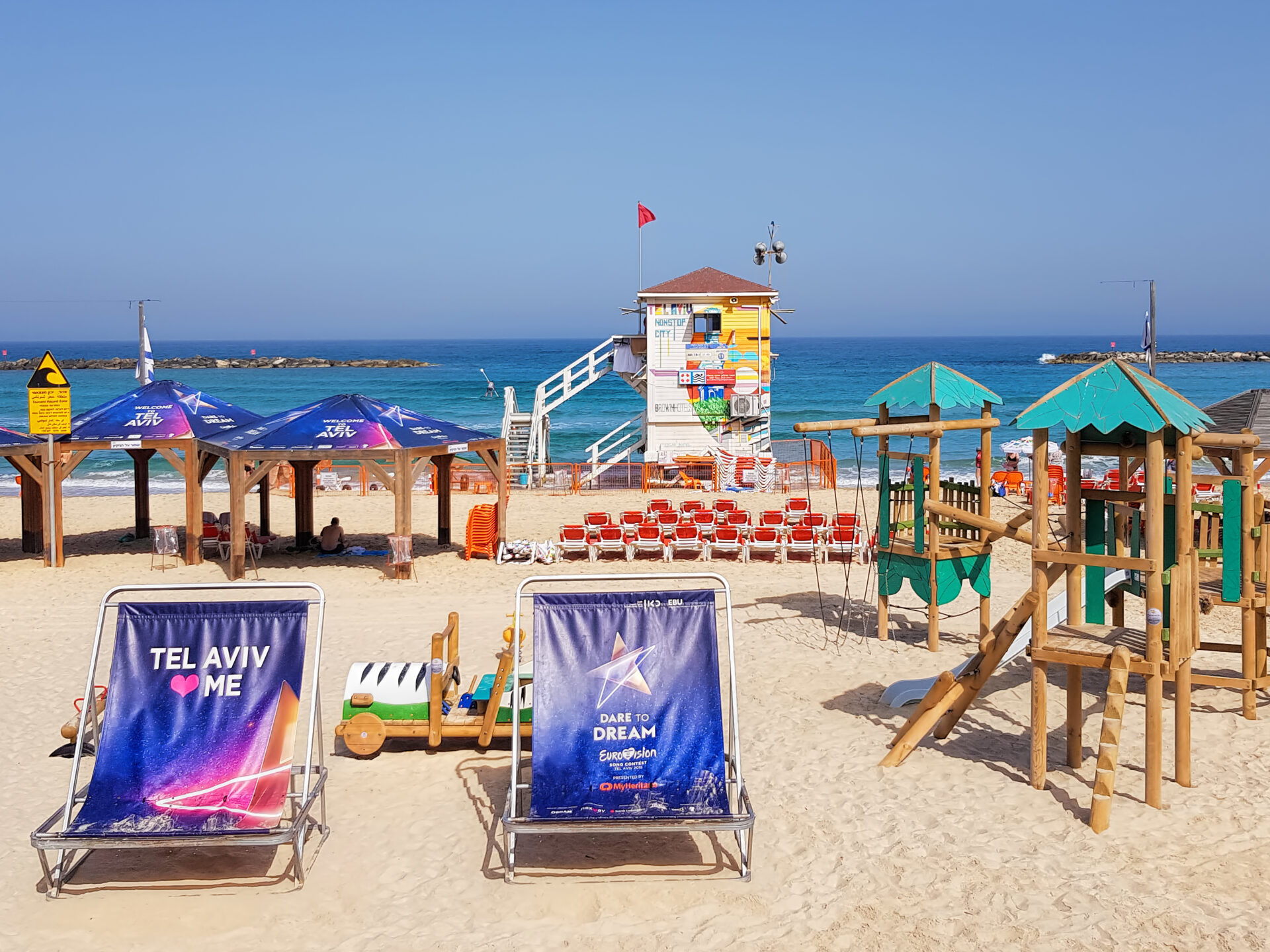 A sunny beach scene on Frishman Beach with a lifeguard tower adorned with various posters in the center. Beach chairs with printed designs and umbrellas are in the foreground. The ocean and clear blue sky are in the background. Theres a play area on the right.