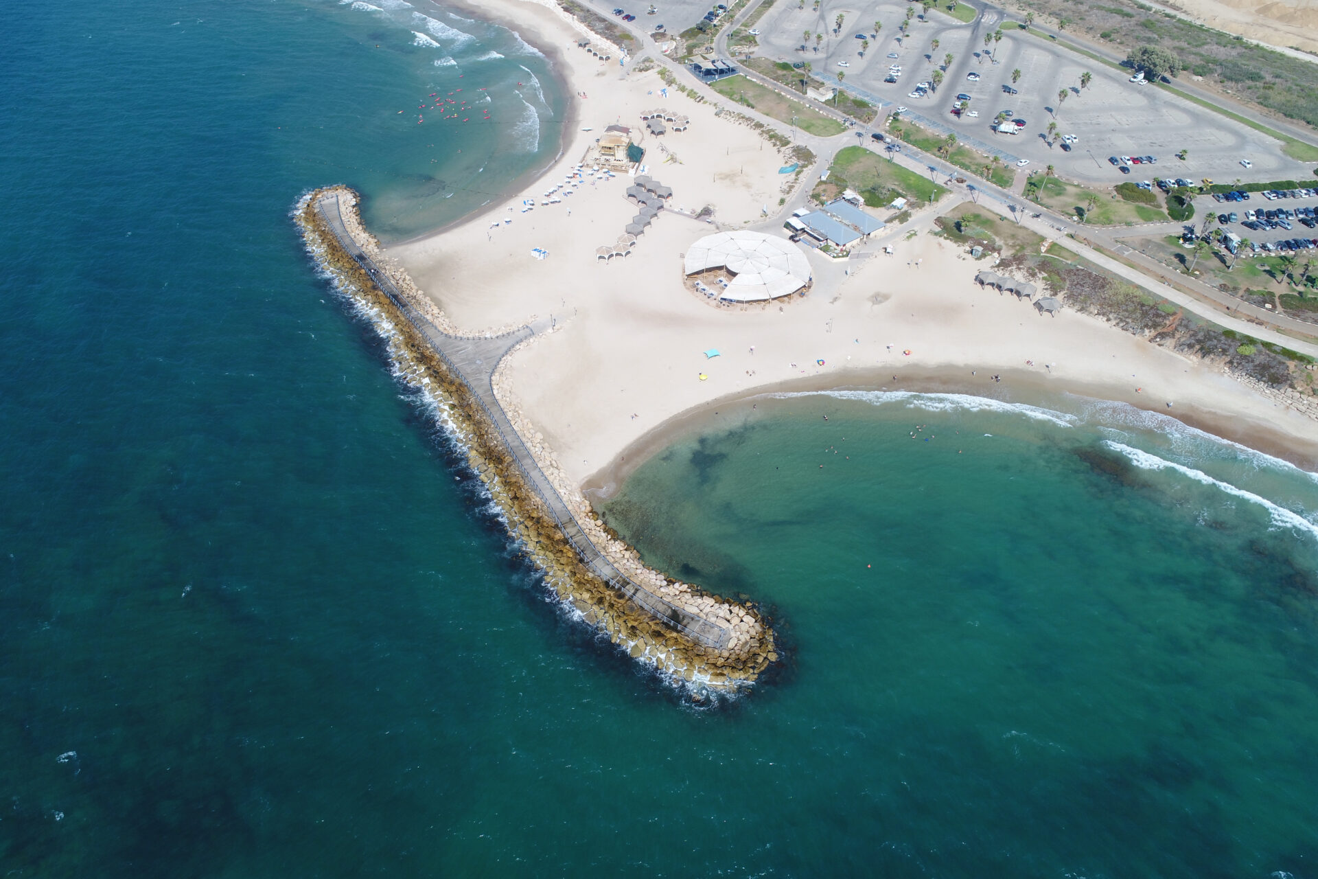 Aerial view of a sandy Tel Baruch beach with a rocky jetty extending into the ocean. A large circular building and parking lot are visible nearby. The sea is deep blue, with waves gently lapping the shore. Sparse beachgoers and umbrellas dot the sand.