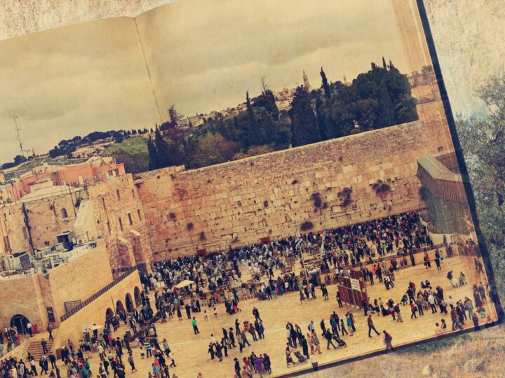 Aerial view of the Western Wall in Jerusalem with a large crowd gathered around. The Dome of the Rock is visible in the background, and the scene is framed as if on an open book page. The sky is overcast, and trees line the horizon.