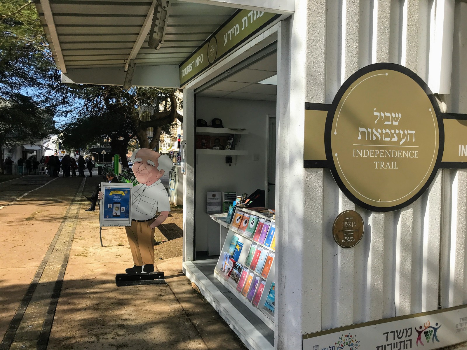 The Independence Trail route guides pedestrians to 10 key sites in Tel Aviv. Photo by Nicky Blackburn Entrance to the Independence Trail in a sunny park, featuring a small kiosk with brochures and books. A cartoon figure Ben Gurion is holding a newspaper near the kiosk. People walk along a path lined with trees in the background.