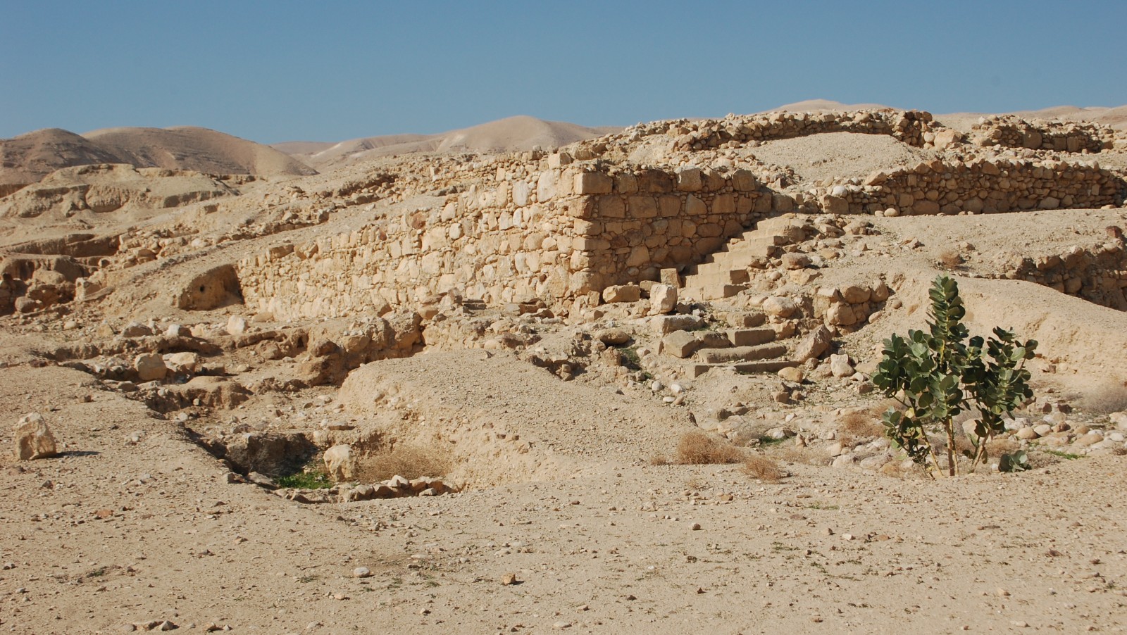 Photo of Hasmonean winter palace ruins by Chaim/Wikimedia Commons Ancient stone ruins in a dry, desert landscape with rocky hills in the background and a few green plants in the foreground under a clear blue sky.