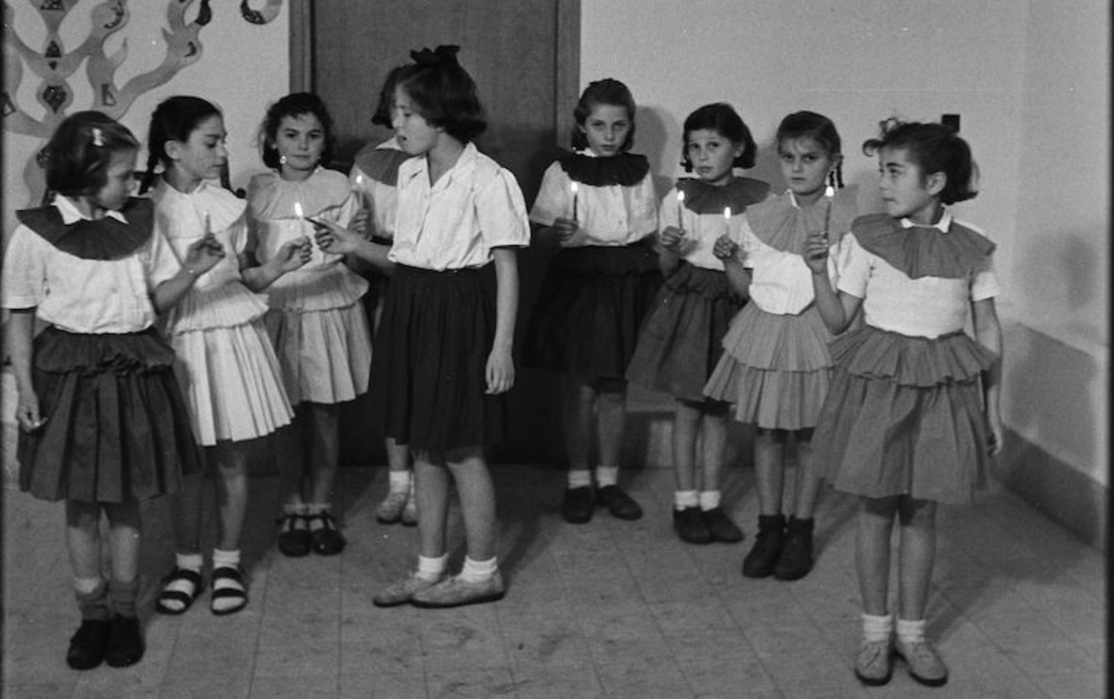A black and white photo featuring a group of seven young girls stand indoors in a row, dressed in matching outfits with short-sleeved tops and skirts, each holding a lit candle. They are participating in a Hanukkah ceremony and celebration.