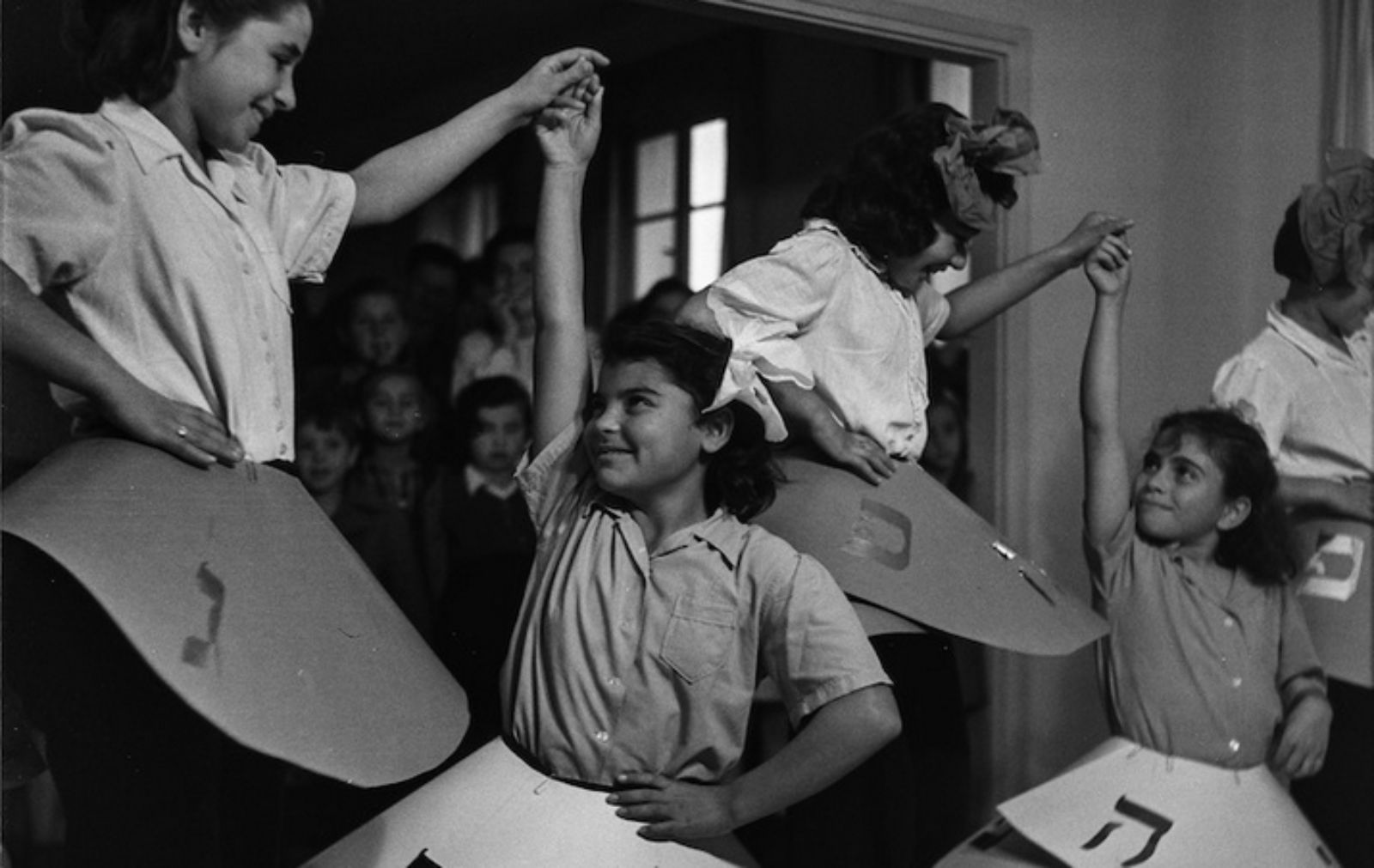 A black and white photo of four young girls dancing indoors wearing large dreidel-shaped costumes, smiling and holding hands, while a group of children watch in the background.