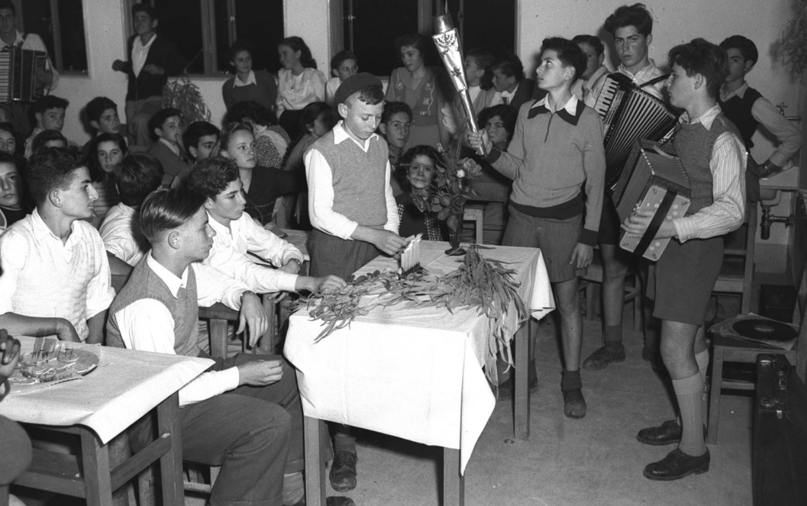 A black and white photo featuring a group of boys and girls sit in a classroom. Some boys stand and play accordion while another holds a bouquet. Several sit around a table with plants and flowers, participating in a Hannukah celebration. 