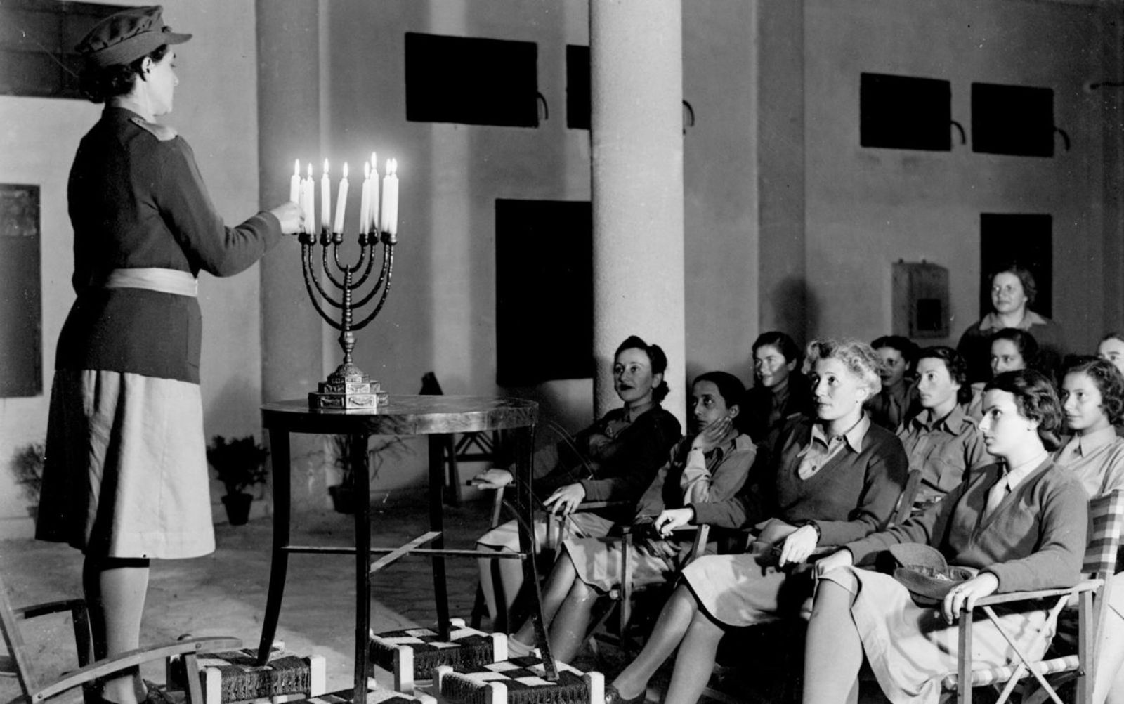 A  black and white photo featuring a woman in uniform who stands beside a menorah with lit candles, leading a Hanukkah candle lighting ceremony for a seated group of women, also in uniform, in a large room with simple decor.