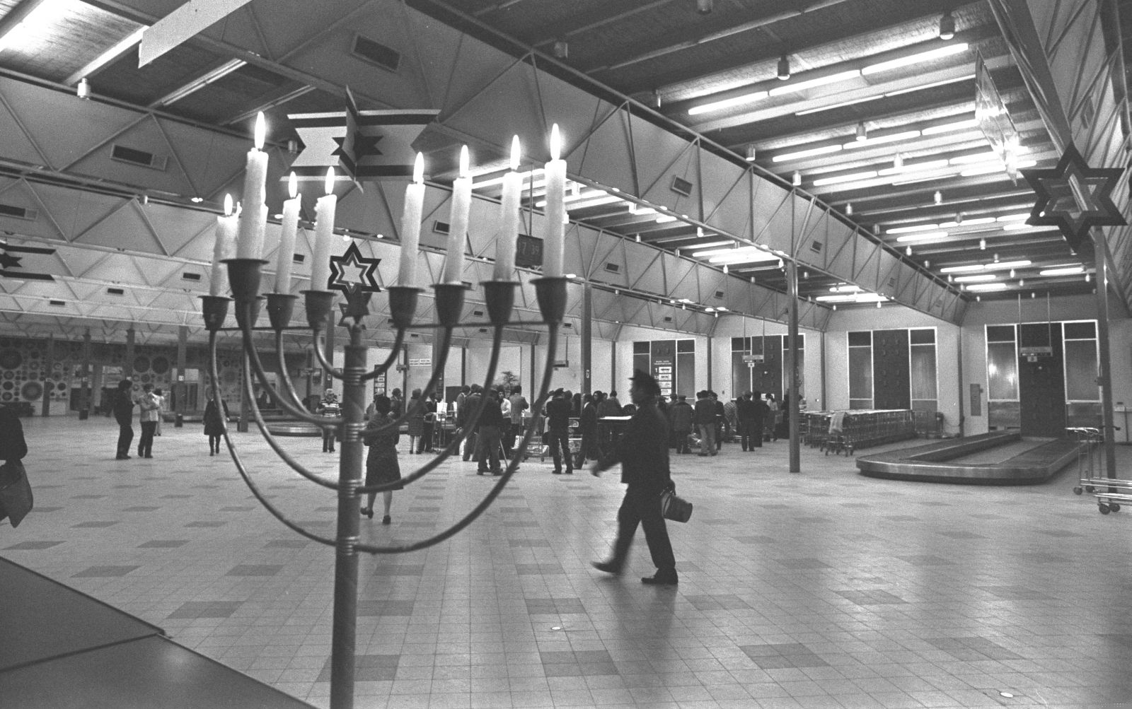 A black and white photo featuring a large menorah with lit candles, stands in the foreground of the spacious, brightly lit hall at Ben Gurion Airport, where people are gathered and walking, some carrying luggage.