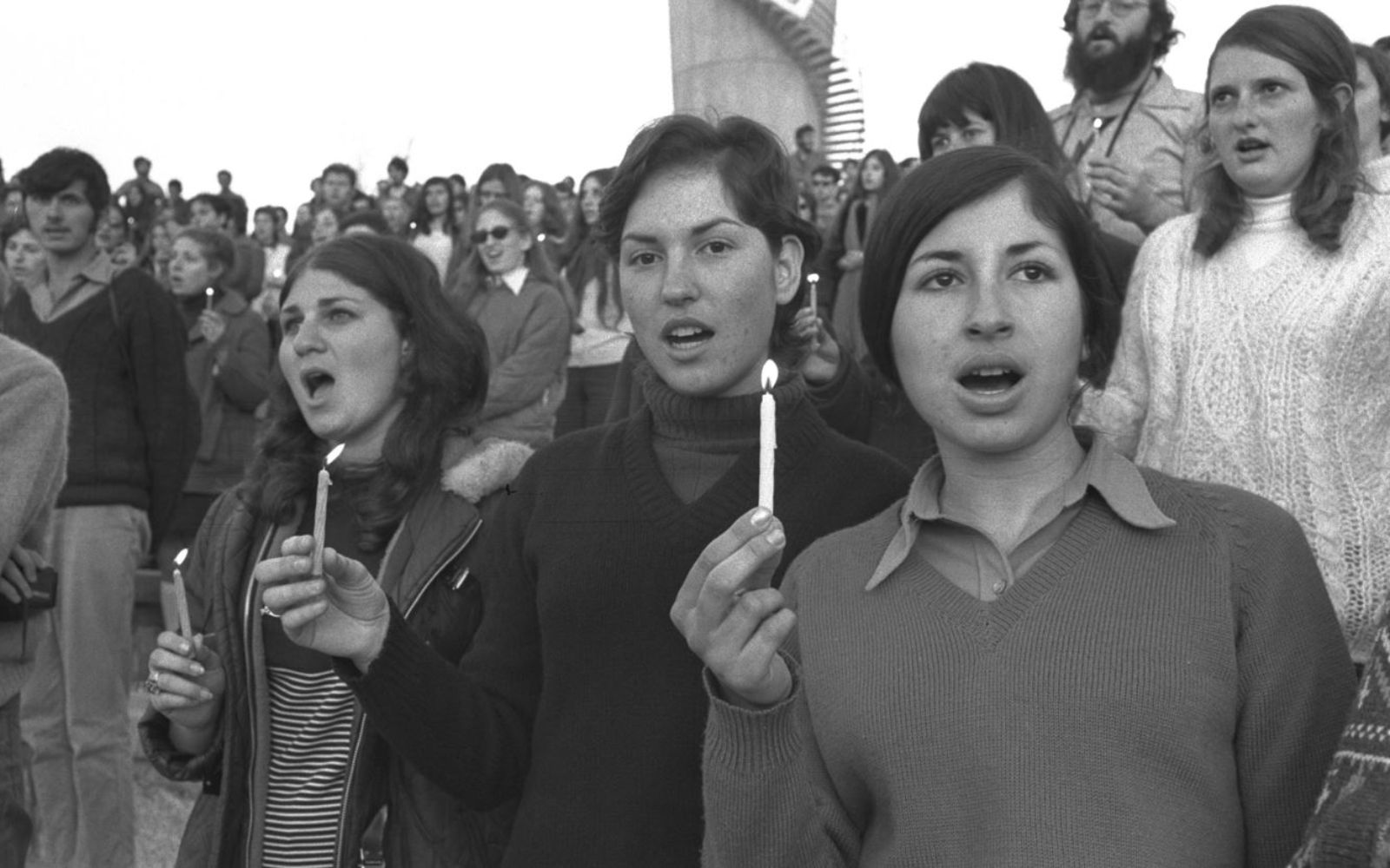 A black and white photo of a group of young  students standing close together, holding lit candles and singing. The scene is set during a Hanukkah lighting ceremony.