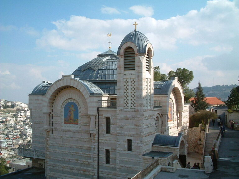 Exterior view of the Church of St. Peter in Gallicantu, featuring its stone facade with a dome and a bell tower, and decorative mosaics and crosses. The building is surrounded by a cityscape under a partly cloudy sky. 