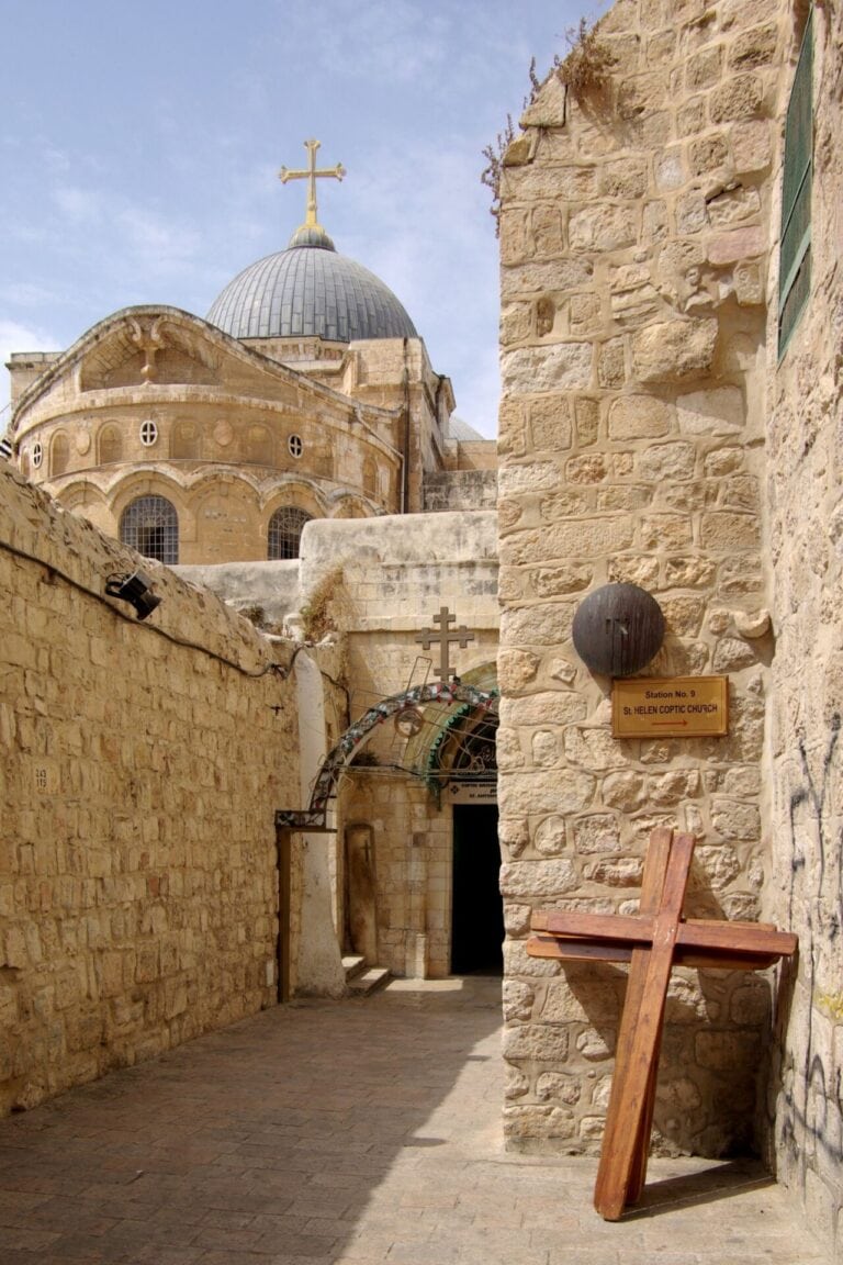 Narrow stone alleyway leading to the historic church of the Holy Sepulchre in Jerusalem. The church dome, topped with a cross, is visible above. Another metal cross and wooden cross are positioned at the entrance with a plaque hung on the wall to the right. The sky in the background is partly cloudy. 