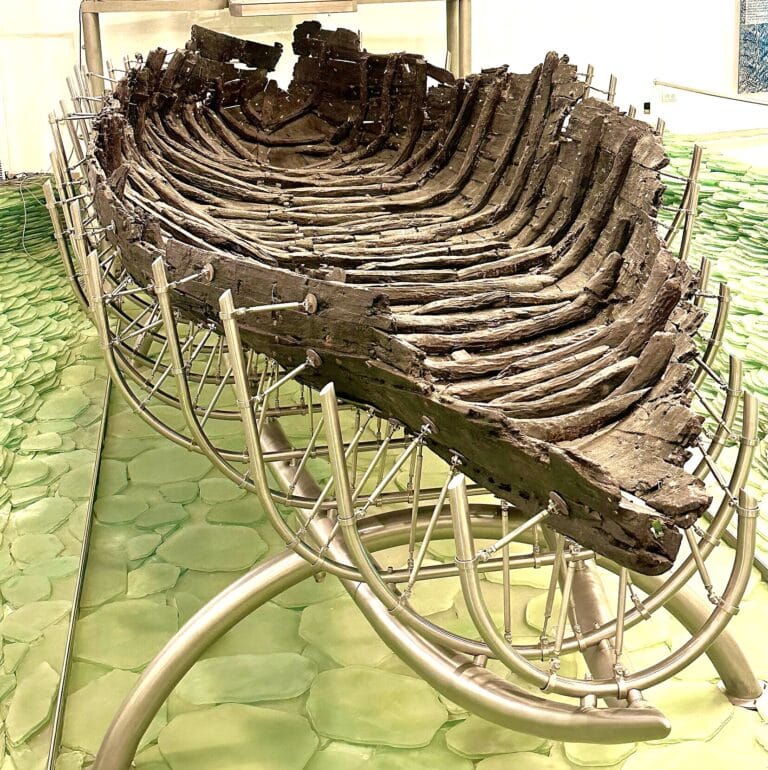 An ancient wooden boat , known as the Jesus Boat, displayed in a museum. The boats structure is supported by a modern metal frame. The background floor is green, resembling a textured surface. The boat appears aged and preserved for exhibition. 