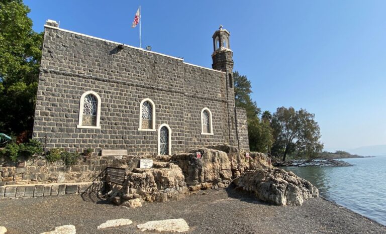 The Church of the Primacy of St. Peter, a dark grey stone building with arched windows and a small tower, is situated near a body of water under a clear blue sky. The building is surrounded by rocks and trees, with a white and red flag flying on top. 