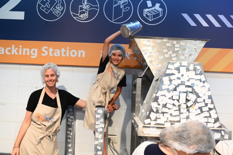 Photograph of a mother on the left and her child on the right, both wearing a hairnet and an apron, inside of a packaging center. The young boy is standing on top of a chair and adding items to a package.