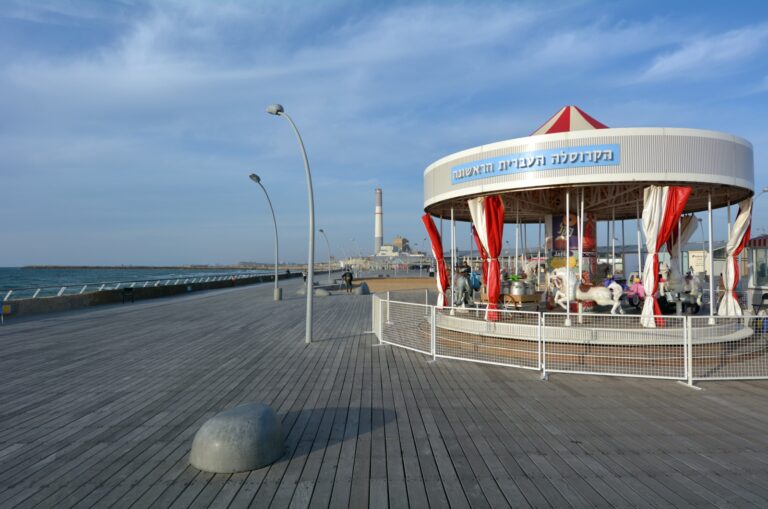 A view of a carousel on the boardwalk of Tel Aviv Port, with a view of the Mediterranean Sea and clear skies in the background.
