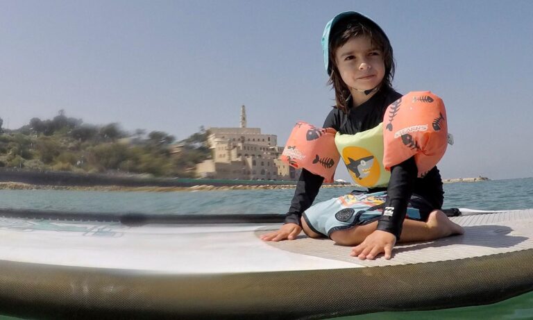 A young girl with floats on her arms and a bucket hat on top of a SUP board in the Mediterranean Sea with Yafo in the background.