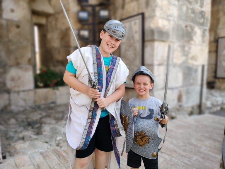 Two young boys wearing knight costumes and holding toy swords smiling outside of the Tower of David museum.