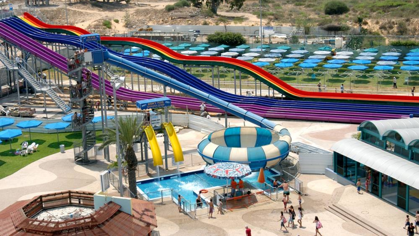 View of Yamit outdoor waterpark features three large waterslides in purple, blue and red colors, two smaller yellow pool slides adjecent, and a handful of visitors walking around.