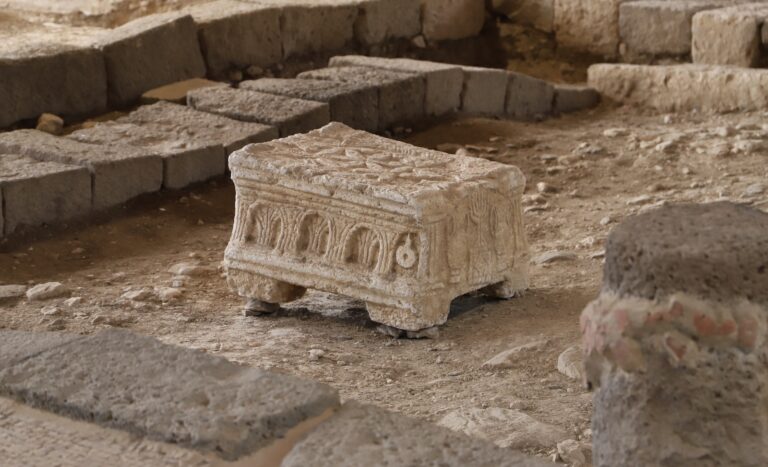 The ancient stone artifact, The Magdala Stone, with intricate carvings, resembling a small pedestal or altar, surrounded by uneven stone floor and partial ruins. The setting appears to be an archaeological site. 