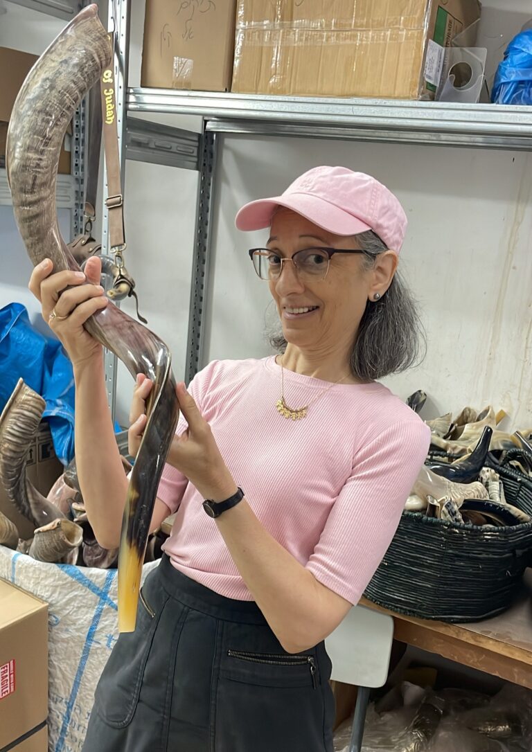 Abigail Klein Leichman holding an antelope horn at Barsheshet & Ribak. Photo by Natalie Selvin A woman wearing a pink shirt and cap smiles while holding a large shofar in a room filled with other shofars and various items on shelves behind her.