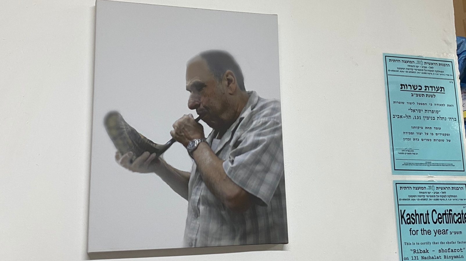 A picture of Eli Ribak’s late father, Avraham, on the wall of Barsheshet & Ribak in Tel Aviv. Photo by Abigail Leichman A photo of man in a plaid shirt blowing a shofar hangs on the wall, with two blue certificates posted on the wall beside him, one in Hebrew and one labeled Kashrut Certificate in English.