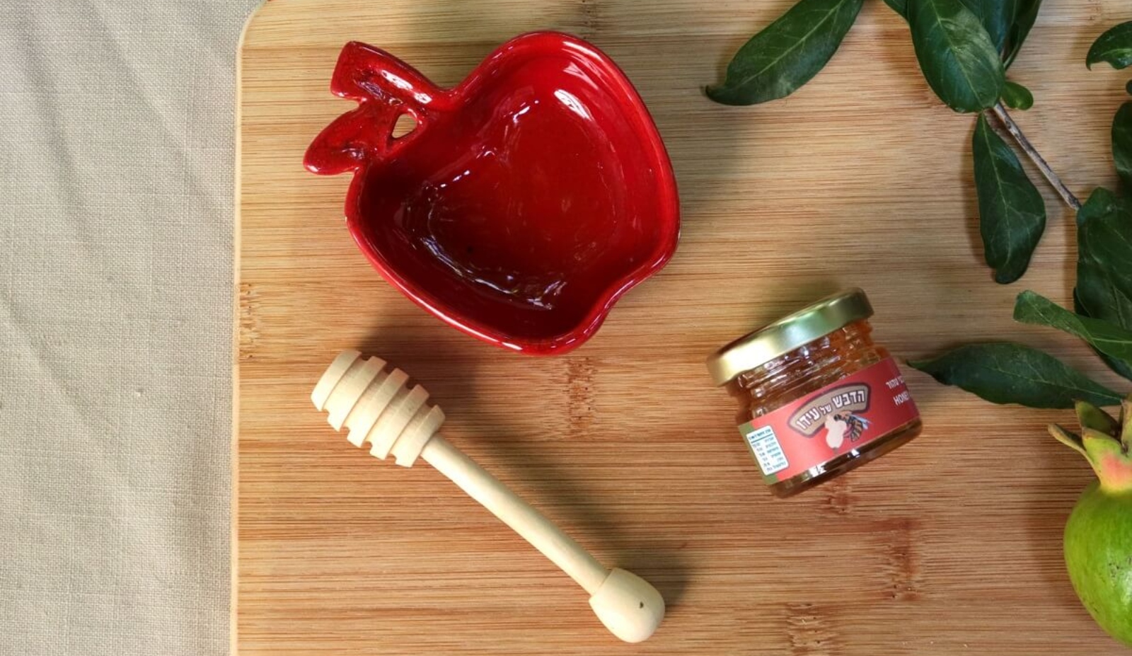 A red apple-shaped dish, a jar of honey, a wooden honey dipper, and green leaves are arranged on a wooden board.