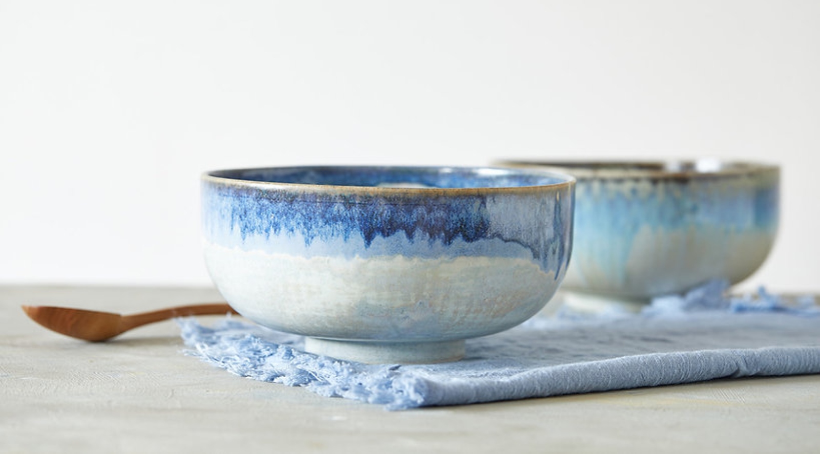 Two ceramic bowls with blue and white glaze sit on a textured blue cloth, with a wooden spoon nearby, against a light background.