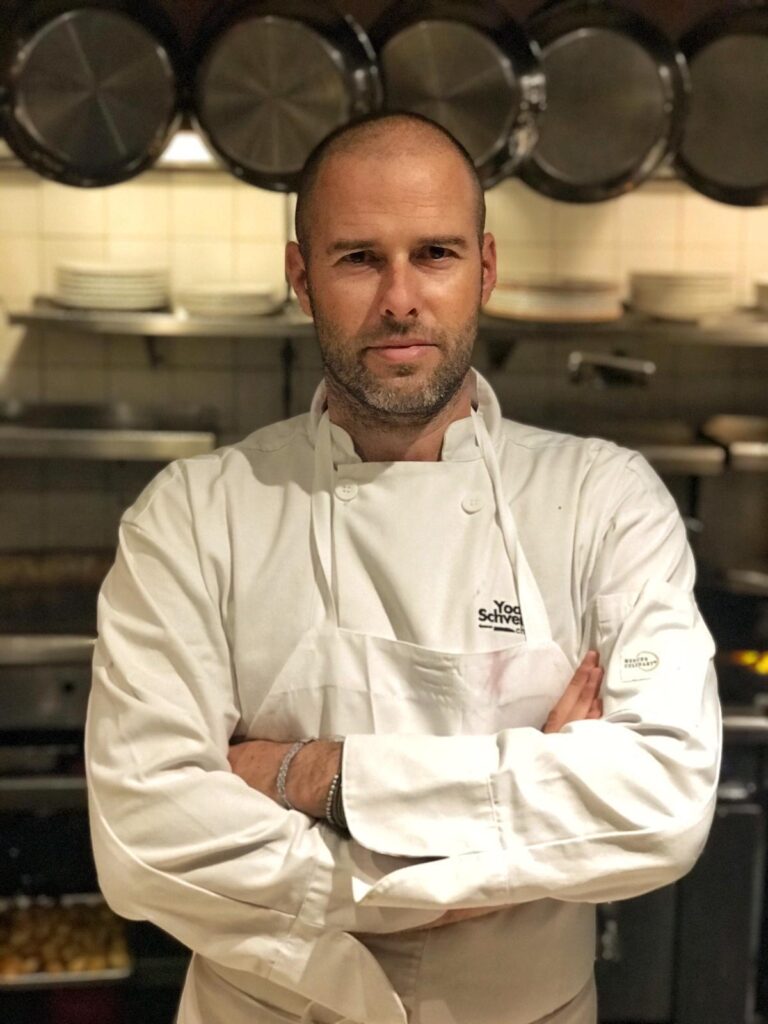 Chef Yoav Schverd with a bald head and stubble stands confidently with arms crossed, wearing a white chefs coat and apron. The background shows a kitchen setting with hanging pans and stacked plates.