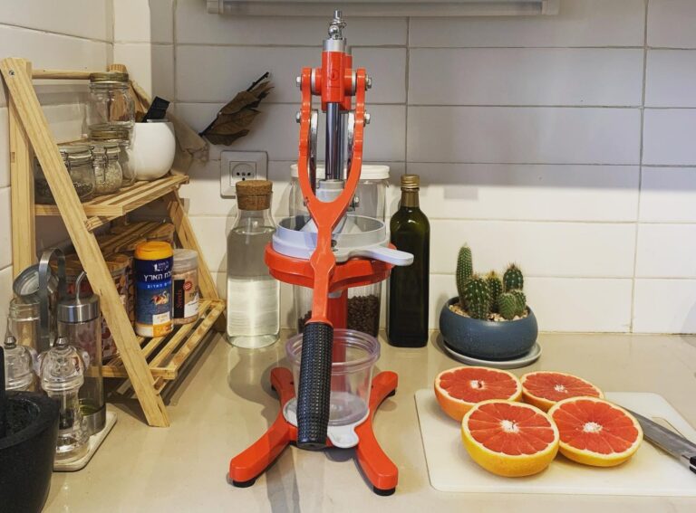 A red manual citrus juicer sits on a kitchen counter next to a bowl of cacti, a shelf with jars and spices, and a cutting board with halved grapefruits ready for juicing.
