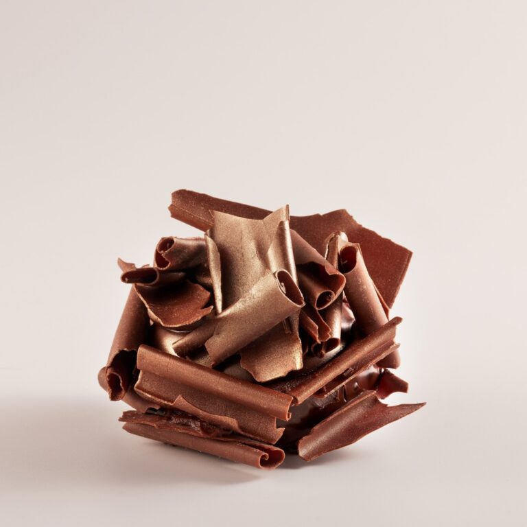 A cluster of curled chocolate shavings, with some pieces coated in a gold metallic finish, arranged against a plain light background.
