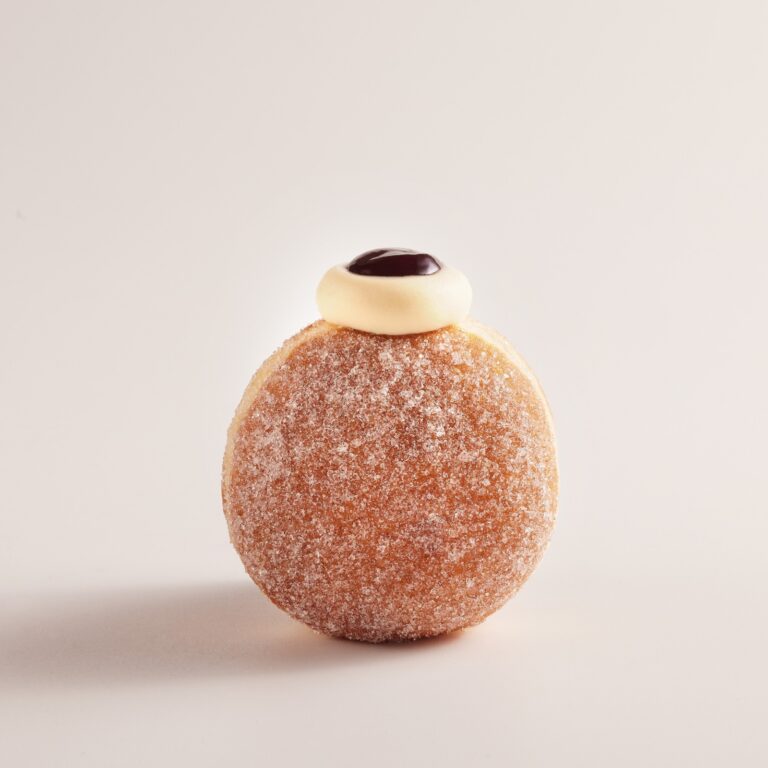 A round sugar-coated doughnut topped with a swirl of white cream and a dollop of dark chocolate or jam, standing upright against a plain light background.
