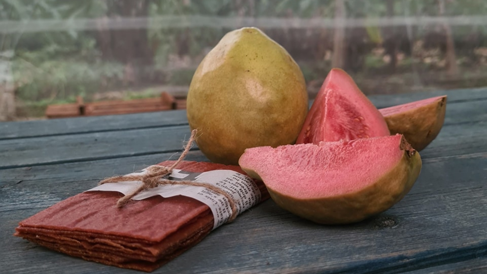 A whole guava and sliced pink guava pieces sit on a wooden surface. Next to them is a stack of dried fruit leather tied with twine. The background is a blurred garden scene. 