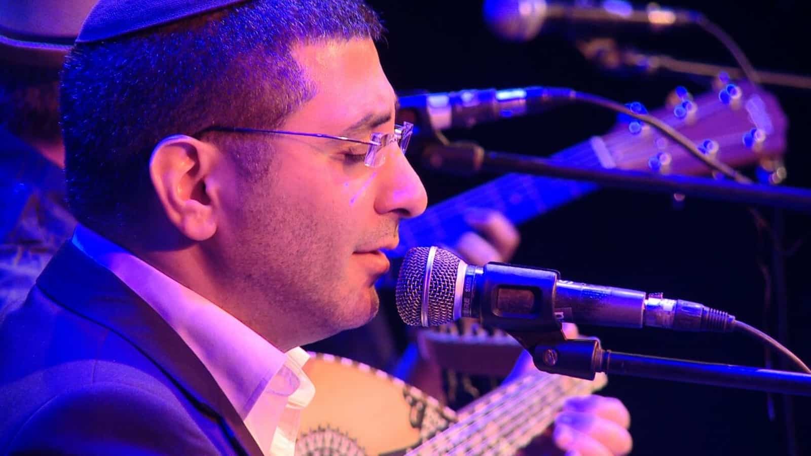 David Menachem, wearing glasses and a kippah sings into a microphone on stage while playing a stringed instrument, with another musician and guitar visible in the background under blue lighting.