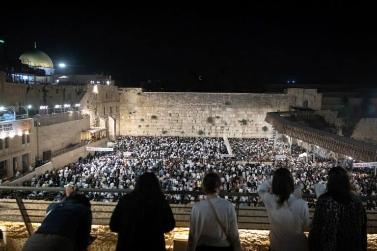 A large crowd gathers at night in front of the Western Wall in Jerusalem for Selichot, while several people observe from a balcony in the foreground.