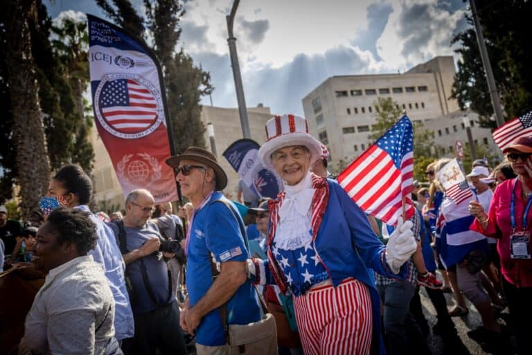 A group of people participate in the Feast of Tabernacles parade in Jerusalem. One person is dressed in an Uncle Sam costume, smiling and holding an American flag. Others around them also wave flags, and trees and buildings are in the background.