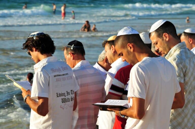 A group of men wearing kippahs stand on the beach near the water, reading from books or prayers, while other people swim in the sea in the background.