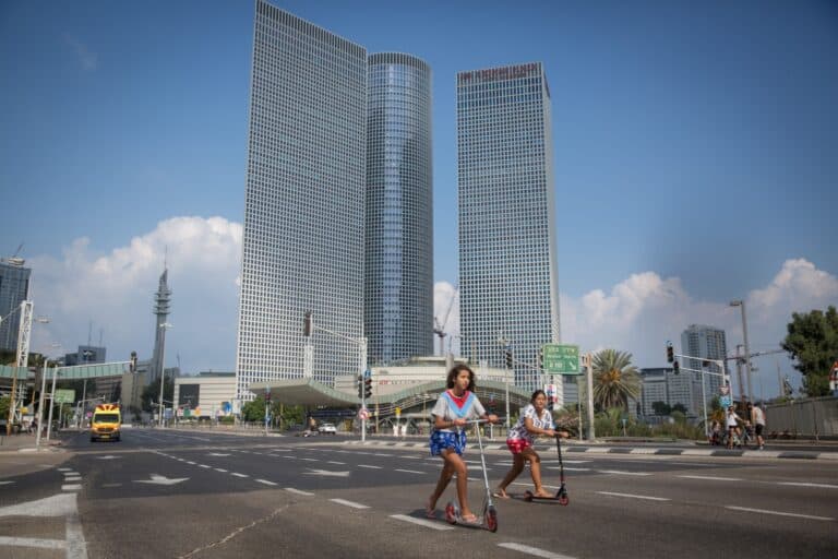Two children ride scooters across a wide and empty city street in Tel Aviv with tall, modern glass skyscrapers and traffic lights in the background on a sunny day.