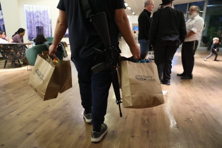 A person carrying a rifle on their back and holding grocery bags in both hands inside a store or mall. Other people are in the background, some seated and others standing on a wooden floor.