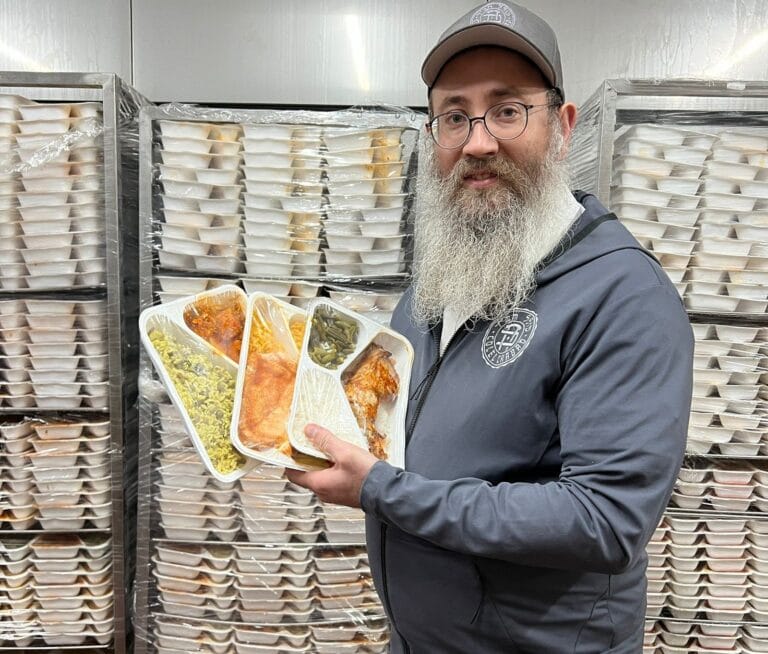 A man with a beard, wearing glasses and a cap, holds packaged meals in a storage room. Numerous trays are stacked on shelves in the background. He is smiling and wearing a dark hoodie.