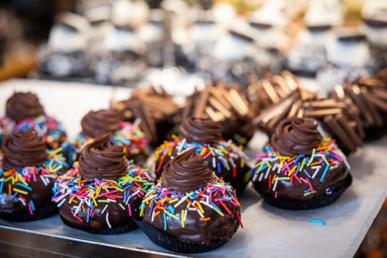 Chocolate cupcakes topped with swirls of chocolate frosting and colorful rainbow sprinkles are displayed on a tray, with more chocolate treats blurred in the background.