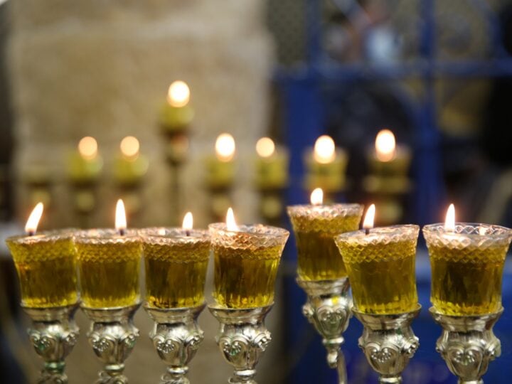 A close-up of a menorah with eight lit oil-filled glass cups, each with a visible flame. Other blurred menorahs are visible in the background.