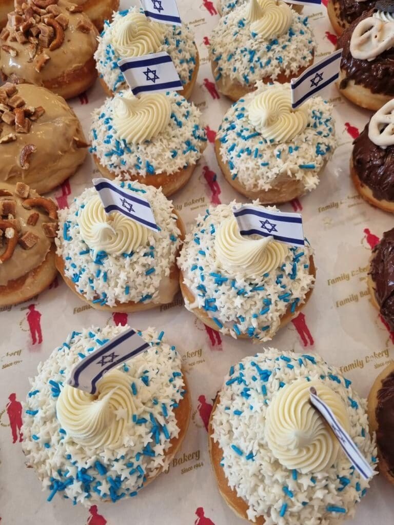 Donuts topped with white icing, blue-and-white sprinkles, piped cream, and small Israeli flags on toothpicks, arranged on bakery paper with a red logo pattern. Other assorted donuts are visible nearby.