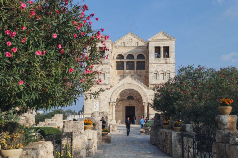 The historic Church of the Transfiguration, with stone, arches and a triangular gable standing under a clear blue sky. Flowering bushes line the stone path leading to the entrance. People are walking near the entrance, enhancing the vibrant, serene scene. 
