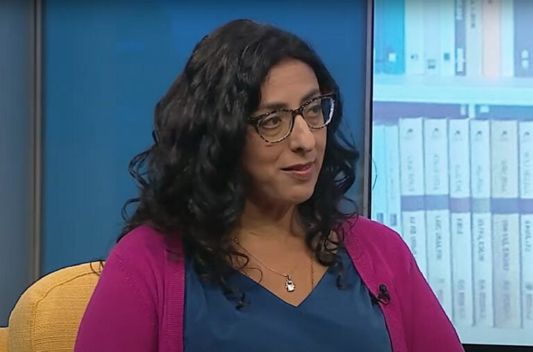 Author Ayelet Tsabari sitting with long curly black hair and glasses. She is wearing a pink cardigan over a blue top. Shelves filled with books are visible in the background. 