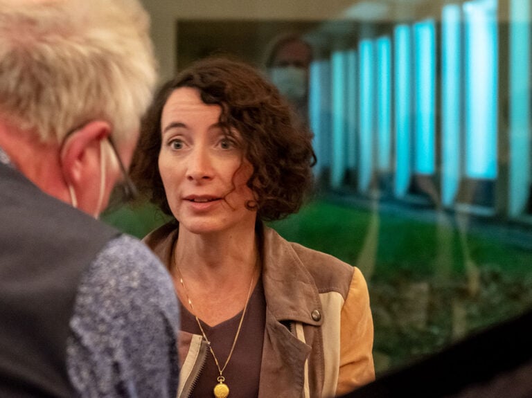 Writer Ayelet Gundar Goshen, with brown curly hair, speaking to a man wearing a mask and glasses. She is indoors, and there is a blurred background with pillars and greenery visible through window reflections. 