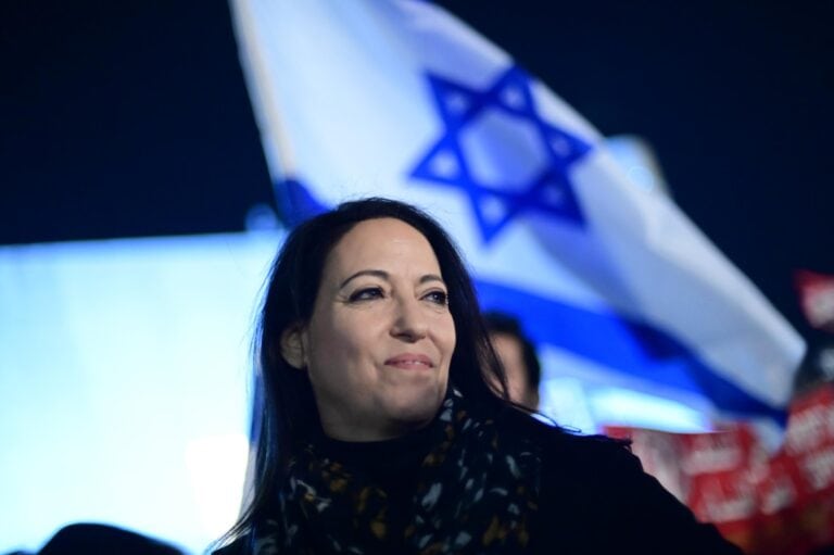 Author Lihi Lapid  smiling with an Israeli flag in the background. She is wearing a dark coat and patterned scarf, and the image is taken at night, giving a dramatic effect with the flag partially illuminated. 