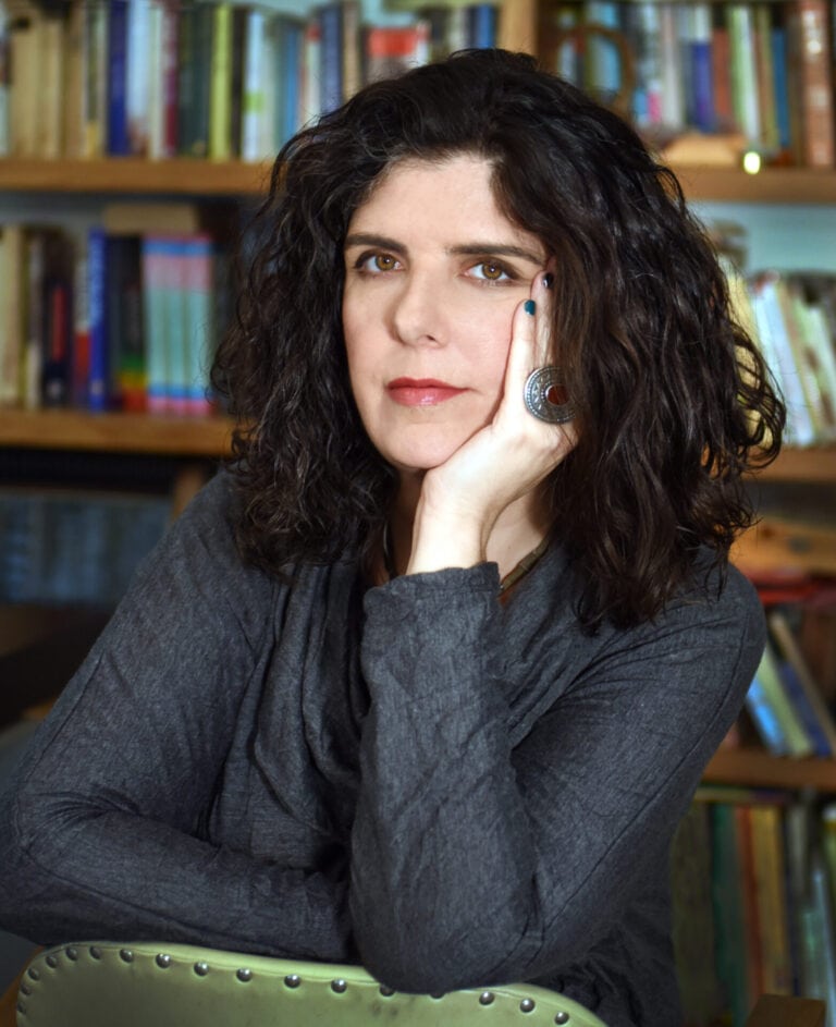 Author Noa Yedlin gazing thoughtfully at the camera, she has long curly dark hair and rests her chin on her hand while seated in front of a bookshelf. She is wearing a grey top and a silver ring. 