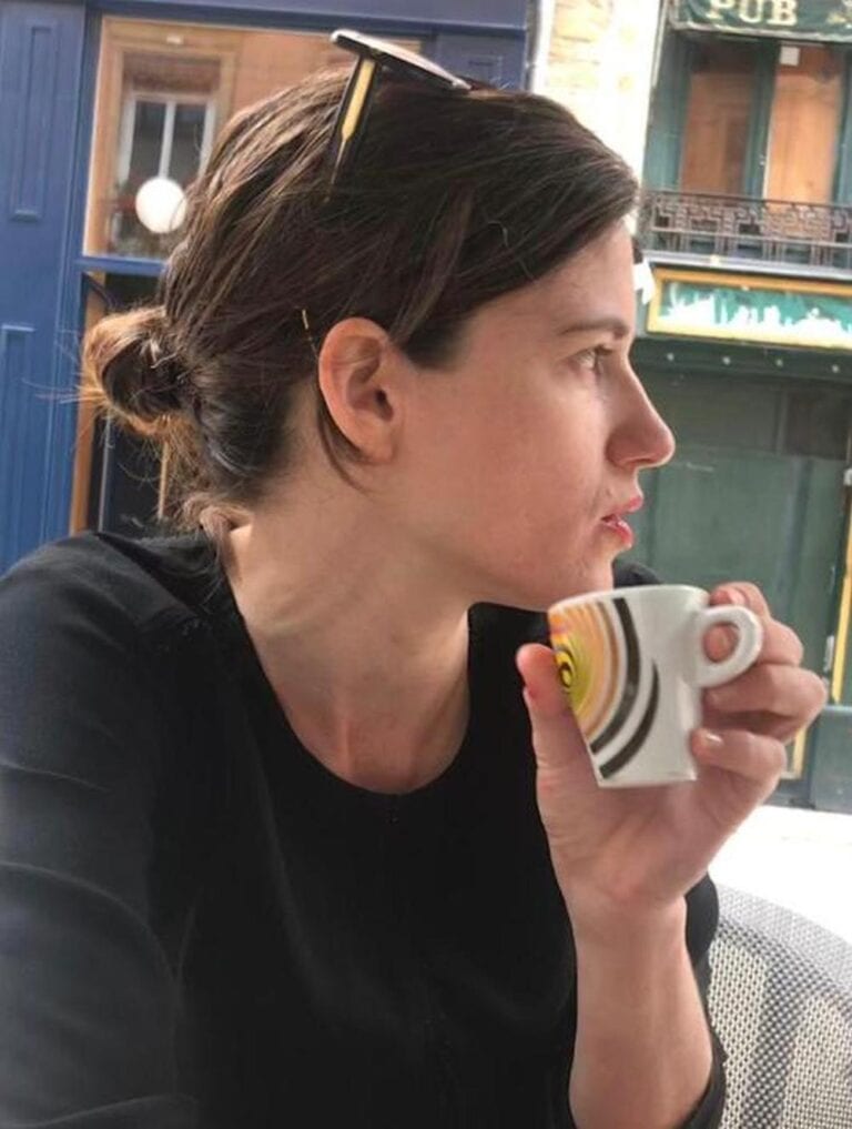 Novelist Yaara Shehori, with dark hair pulled back and sunglasses on her head, sipping from a small espresso cup. She is wearing a black top and is seated outdoors at a cafe, gazing to her right. The background shows a blurred street scene with blue and wooden tones. 