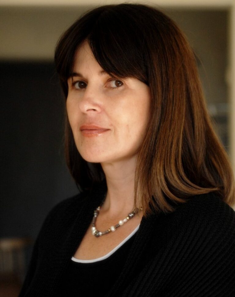 Novelist Yochi Brandes with shoulder-length brown hair, waring a black sweater over a white top and a beaded necklace, looking at the camera with a neutral expression. The background is softly blurred. 