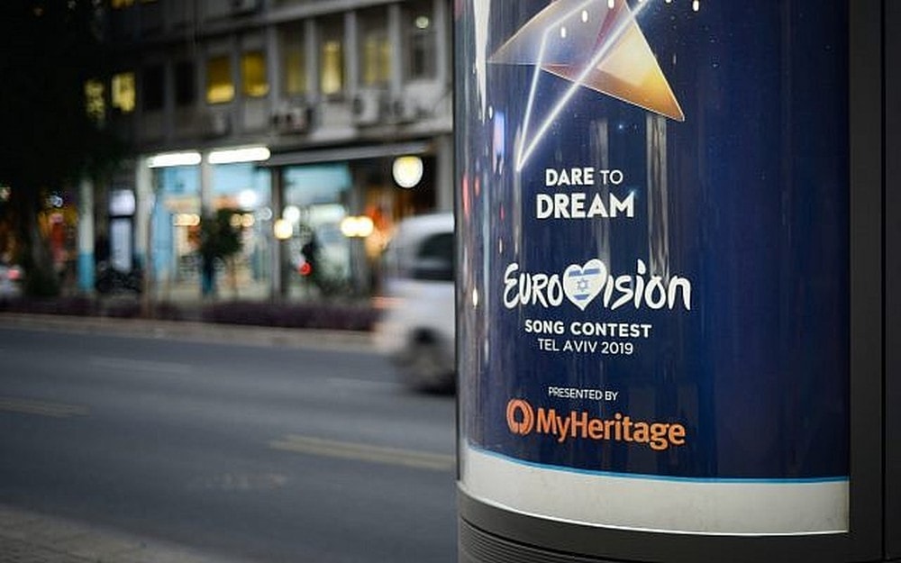 A street advertisement displays the Eurovision Song Contest 2019 poster with the slogan “Dare to Dream,” held in Tel Aviv, and presented by MyHeritage. Cars and buildings are visible in the background.